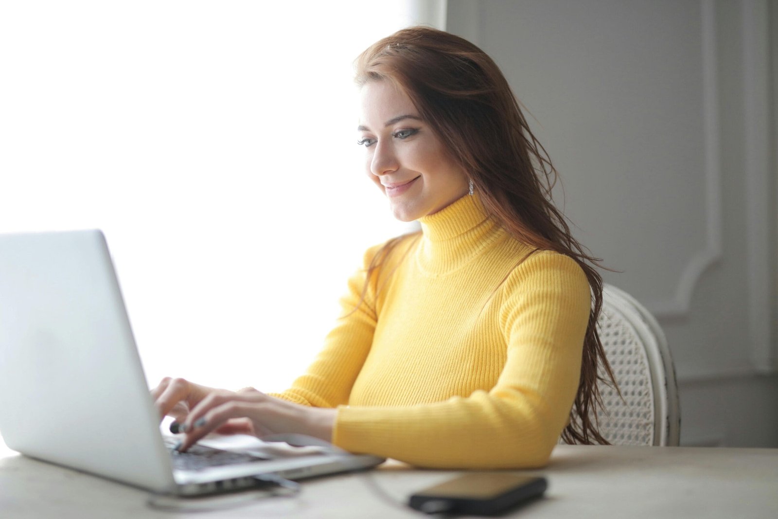 Smiling woman in yellow sweater working on laptop indoors, focused and engaged.