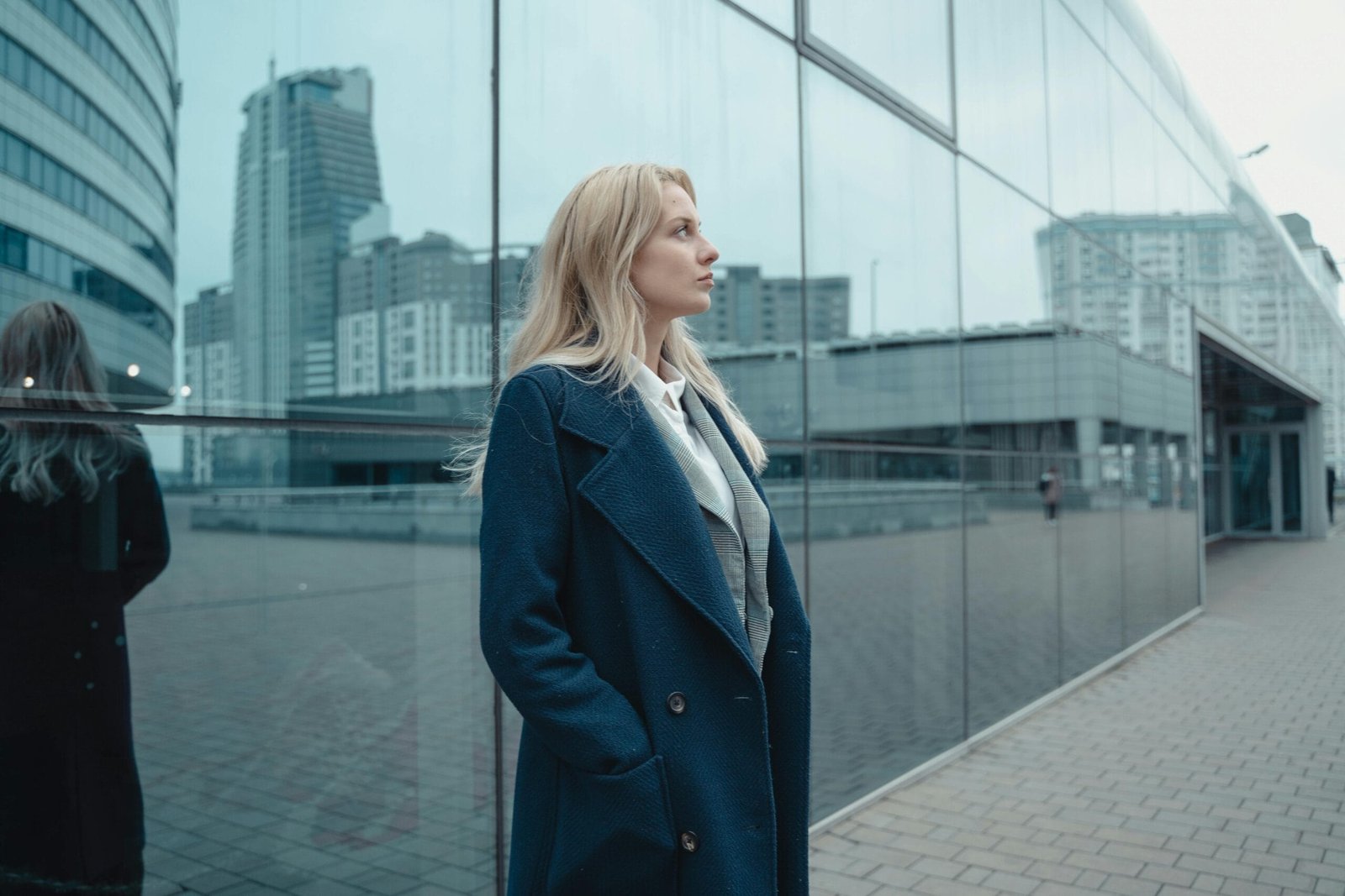 A businesswoman in a coat stands confidently against a glass building with city skyscrapers.