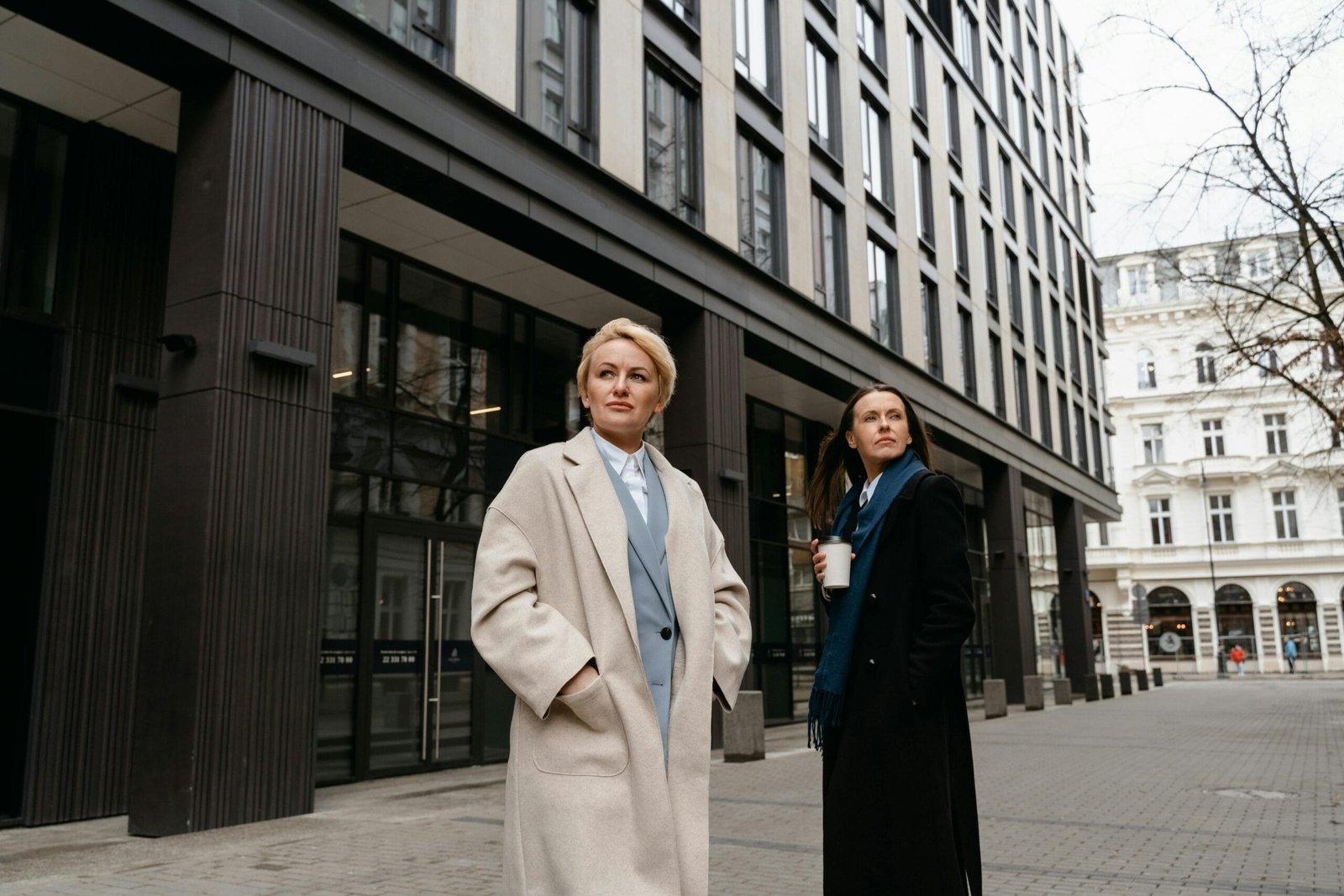 Two women in stylish overcoats walk outside modern office buildings.